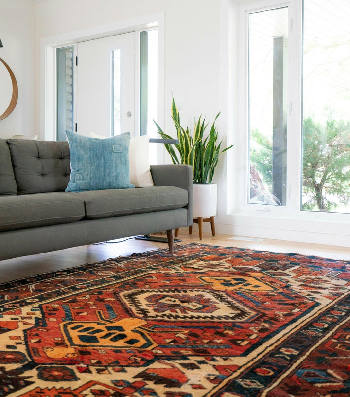 Showcasing a modern living room with a minimalist style. It features a grey mid-century modern sofa with tufted cushions. The sofa is placed on a vibrant, intricately patterned Turkish rug dominated by shades of red, blue and beige. 